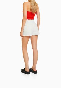 Top corto fruncido rojo combinado con pantalones cortos de mezclilla blancos. Los pantalones cortos tienen un dobladillo deshilachado y dos bolsillos traseros, complementados con sandalias negras de plataforma.