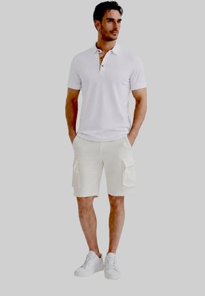 Uomo che indossa una polo bianca a maniche corte, pantaloncini cargo bianchi e sneakers bianche, in piedi con le mani in tasca su sfondo grigio.