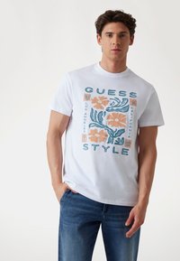 Witte katoenen t-shirt met een blauw en oranje bloemen grafisch ontwerp, met de tekst "GUESS" en "STYLE" in vetgedrukte letters.