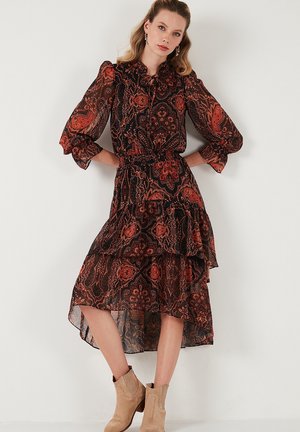 Long, flowy dress in black fabric with red paisley pattern, ruffled hem, puffed sleeves, and a tie-neck detail. Paired with beige ankle boots.