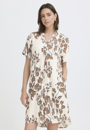 Femme portant une robe crème à manches courtes avec des motifs floraux abstraits marron et noir, debout devant un fond clair uni.