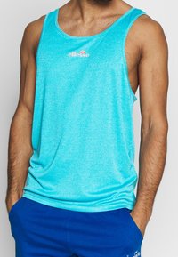 Uomo che indossa un top sportivo senza maniche di un brillante blu con il logo "ellesse" e pantaloncini blu coordinati, mani in tasca, su sfondo neutro.
