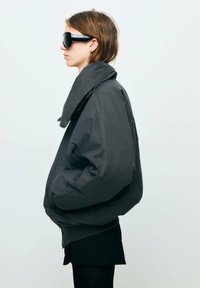 Przesadnie duża czarna kurtka z wysokim kołnierzem, dużymi kieszeniami i teksturowanym wykończeniem, noszona z dopasowaną czarną spódnicą i ciemnymi okularami przeciwsłonecznymi.