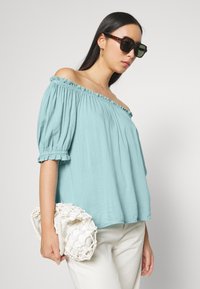 Blusa azul clara sem alças com mangas bufantes, apresentando bordas franzidas. Modelo segura uma clutch branca texturizada.