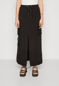 Falda negra hasta el tobillo con abertura frontal, dobladillo ajustable con cordón, bolsillos laterales tipo cargo con malla, combinada con sandalias de tacón bloque negro con tiras.