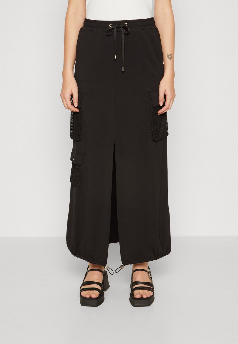 Falda negra hasta el tobillo con abertura frontal, dobladillo ajustable con cordón, bolsillos laterales tipo cargo con malla, combinada con sandalias de tacón bloque negro con tiras.