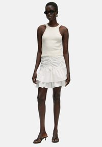 Witte geribbelde haltertop gecombineerd met een gelaagde witte minirok, met ruchingdetails, gedragen met zwarte zonnebril en minimalistische sandalen.