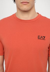 Rød bomull T-skjorte med rund hals, med en svart EA7-logo på venstre bryst og korte ermer. Ensfarget, glatt tekstur.