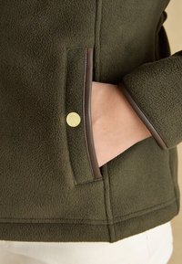 Hand in olive green fleece jacket pocket with brown leather trim and gold snap button, showing jacket texture and cuff detail.