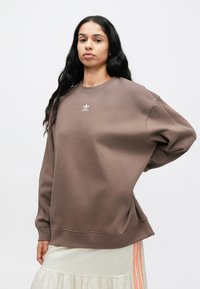 Καφέ φούτερ με άνετη γραμμή, στρογγυλή λαιμόκοψη και κεντημένο λευκό λογότυπο. Απαλό μείγμα βαμβακιού, με ριχτούς ώμους και ριμπ τελειώματα στα μανίκια.