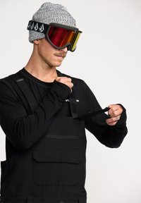 Zwarte snowboard overalls met verstelbare banden, voorzien van een voorzak en een aansluitend zwart longsleeve t-shirt. Grijze gebreide beanie en kleurrijke skibril.