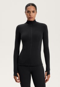 Giacca sportiva nera con maniche lunghe, colletto alto e chiusura con zip; presenta un tessuto testurizzato e aperture per il pollice sui polsini.