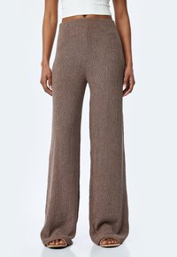 Pantalons amples marron en tissu texturé, dotés d'une taille haute et d'une finition lisse, associés à des chaussures ouvertes.