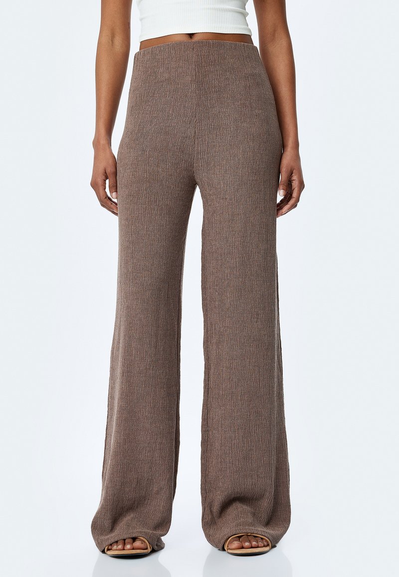 Pantalons amples marron en tissu texturé, dotés d'une taille haute et d'une finition lisse, associés à des chaussures ouvertes.