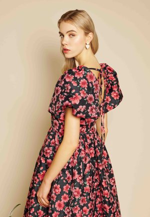 Ung kvinde med langt blondt hår iført sort kjole med rød blomsterprint og pufærmer, vendt så ryggen vises bundet med sløjfe.