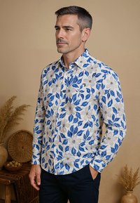 Witte blouse met blauwe bloemen- en bladerenpatronen, met knopen en lange mouwen. Gekozen over donkere broek, met een effen achtergrond.