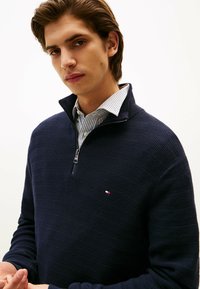 Pullover blu navy lavorato a maglia con zip a un quarto, caratterizzato da strisce orizzontali texturizzate e un piccolo logo sul petto. Indossato sopra una camicia con colletto.