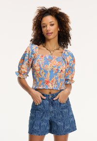 Kvetinový crop top s puff rukávmi v modrej farbe, zdobený riasenou textúrou a farebnými kvetinovými vzormi. Skombinovaný s džínovými šortkami s vysokým pásom.