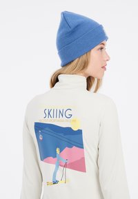 Crème kleurige longsleeve top met hoge hals en een kleurraf ski-ontwerp op de achterkant; een blauwe gebreide beanie complementeert de outfit.
