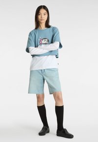 Blå cropped t-shirt med Vans-logo, lagdelt over en hvid langærmet skjorte. Lyseblå denimshorts, sorte knæhøje strømper og sorte slip-on sko.