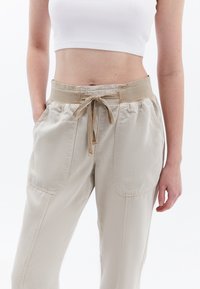 Lichtbeige broek van zachte stof met een elastische tailleband, koord en twee zijzakken, met een losse, ontspannen pasvorm.