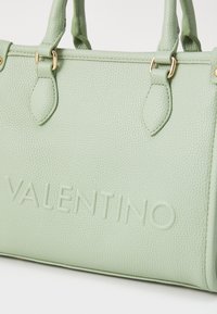 Zelena teksturirana usnjena torba z dvema ročajema, zlato-tonirana kovinska oprema in odtisnjenim logotipom "VALENTINO" na eni strani.