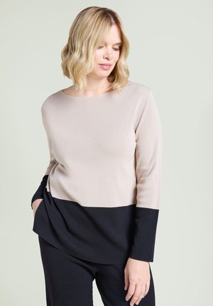 Kvinna med blont hår som bär en beige och svart långärmad topp och svarta byxor, tittar nedåt med ena handen i fickan.