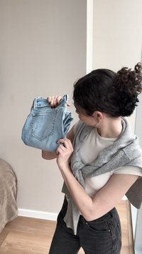 Mujer con el pelo rizado recogido, vistiendo una camisa blanca y un suéter gris, sosteniendo y examinando unos vaqueros azul claro doblados en interiores.
