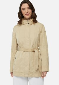 Giacca beige chiaro con cintura, colletto alto, chiusura con bottoni a pressione, tasche laterali e dettagli testurizzati sulle maniche. Tessuto liscio e leggero.