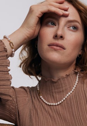 Szereg białych pereł z złotą zapięciem, noszona jako naszyjnik, w połączeniu z pasującą bransoletką z pereł. Teksturalny brązowy top w prążki.