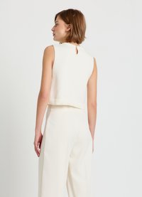 Gekürzter, ärmelloser Pullover aus cremeweißem Strick mit kleinem Rückenschlitz und geripptem Bündchen, kombiniert mit hellfarbigen Hosen.