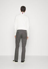 Camicia bianca a maniche lunghe con colletto, con righe rosse e blu, abbinata a pantaloni grigi e sneakers nere su uno sfondo semplice.