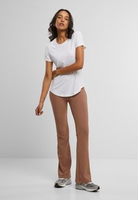 Witte t-shirt met korte mouwen en een ronde zoom, gecombineerd met bruine flare broek. Het model draagt grijze sportschoenen en staat voor een lichte achtergrond.