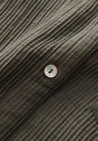 Tissu vert olive texturé avec des rainures verticales, présentant un bouton rond et brillant en nacre avec un seul boutonnière.