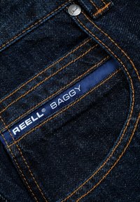Dark denim jeans featuring orange stitching, a tag reading "REELL BAGGY," and a metal button. Close-up of back pocket details.