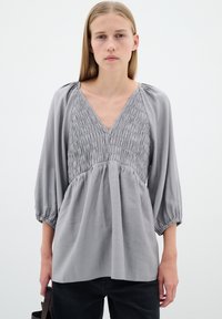 Hallitusvärvi pluus, millel on kokku volditud rinnaosa ja V-kaelus. Omab laiu varrukaid, mis on kogutud varrukate otstes, ning lahedat, voolavat disaini.