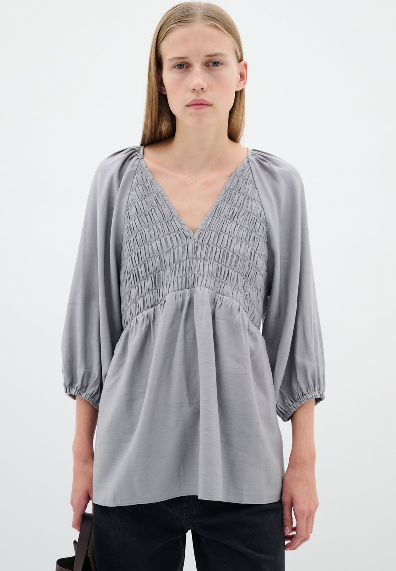 Hallitusvärvi pluus, millel on kokku volditud rinnaosa ja V-kaelus. Omab laiu varrukaid, mis on kogutud varrukate otstes, ning lahedat, voolavat disaini.