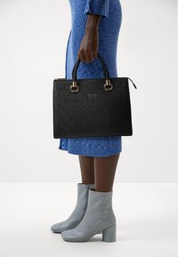 Fekete texturált kézitáska aranyszínű kiegészítőkkel, két felső fogantyúval és oldalsó cipzárral. Egy kék bordás ruhával és szürke bokacsizmával viselve.