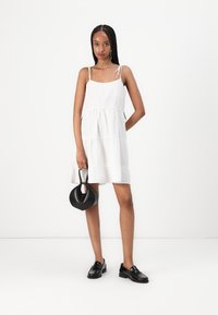 White tiered dress with thin straps, smooth fabric, and a relaxed silhouette. Paired with black loafers and a small black handbag.