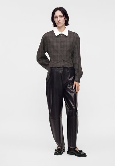 Femme aux cheveux courts et foncés portant une veste foncée à carreaux avec un col blanc, un pantalon en cuir noir taille haute et des mocassins noirs, debout devant un fond blanc.