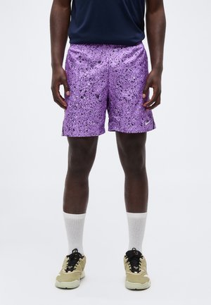 Mand iført lilla atletikshorts med pletter, hvide crew-sokker og beige sneakers står mod en hvid baggrund.