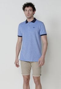 Gai zila polo krekls ar tekstūru, tumši zils apkakles un piedurkņu maliņas, trīs pogu aizdare un izšūta logo. Apvienots ar bēša šortiem.