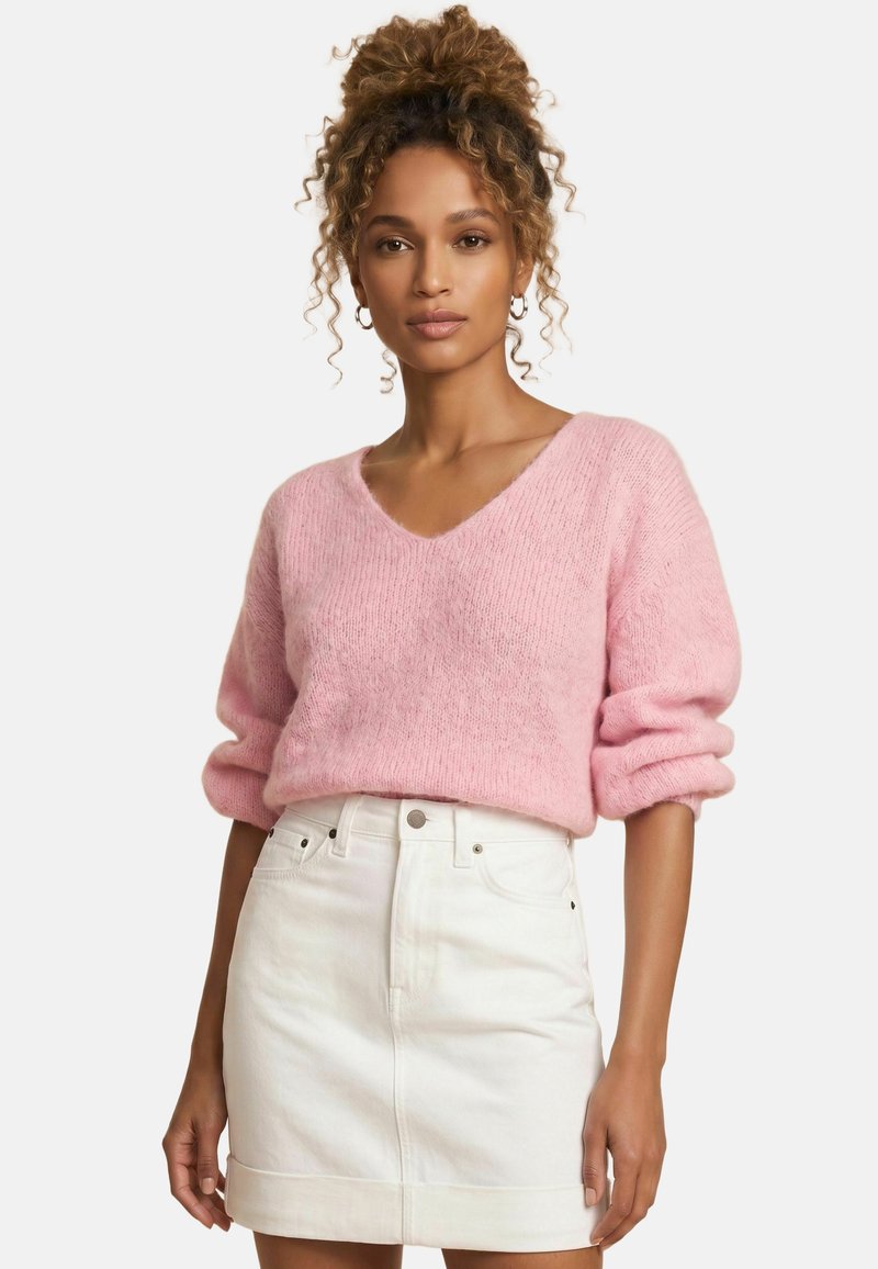 Femme debout, portant un pull rose pâle à col en V et manches longues et une mini-jupe blanche en jean taille haute sur fond uni.