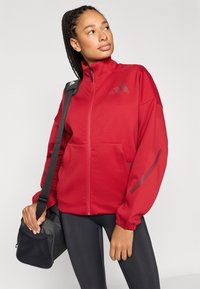 Červená bunda na zips s vysokým golierom, elastickými manžetami a bočnými vreckami. Na hrudi a rukávoch sú jemné grafické detaily.