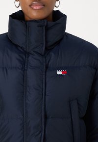 Tumši zili pūkainais jaka ar augstu apkakli, horizontālām šuvēm un izšutu logo sarkanos, baltos un zilos toņos uz kreisā krūtīm. Gluda tekstūra.