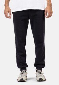 Sorte sweats lavet af en bomuldsblanding, med et tapered design, ribbeede manchetter og sidelommer. Båret med grå træningssko.