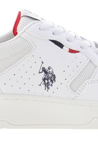 Sneaker bianca con una tomaia in pelle testurizzata, dettagli in rete, perforazioni e una scena stampata di polo sul lato. Dettagli sottili in rosso e blu.