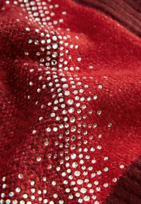 Červená látka s texturovaným povrchem, zdobená vzorem z jasných rhinestones, vytvářející třpytivý efekt na materiálu.