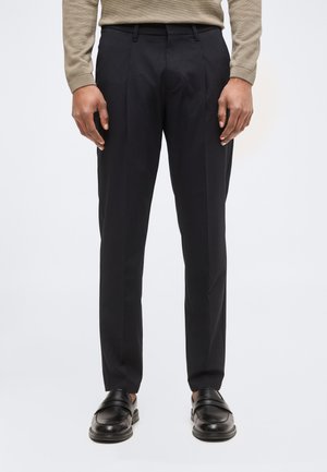 Mann trägt schwarze maßgeschneiderte Hose, schwarze Lederloafers und ein beiges Langarmhemd, steht vor einem einfarbigen Hintergrund.