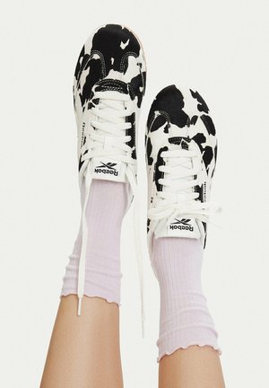 Pies que llevan zapatillas Reebok con estampado de vaca en blanco y negro, cordones blancos y calcetines acanalados rosa claro sobre un fondo liso.